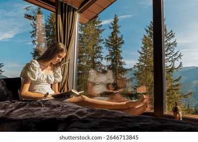 Girl reading by cabin window with mountain view. Teen girl relaxes on a bed, reading a book beside a large glass window with scenic forest and mountain views outside.