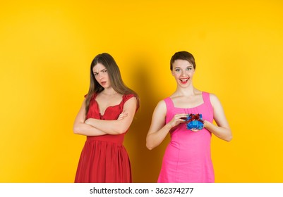 The girl received a gift, and the other girl looks with angry. Isolated studio  yellow background female model.