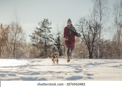 Girl In A Red Down Jacket On Walk With Purebred Beagle Puppy In Winter Park. Woman And Dog In Forest. Girl Runs And Plays In The Snow With Pet. Winter Sunny Landscape. Stylish Toning And Soft Focus.