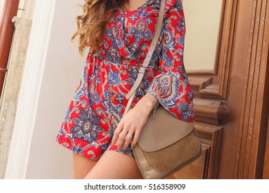 Girl in red dress with beige leather handbag on the shoulder. Woman in summer overall in the city on sunny summer day. Vogue woman with long blonde hair near wooden door