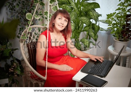 A girl in red dress with a laptop is working in chair at a table. A middle-aged business woman. Funny psychologist in the office with flowers