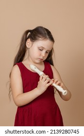 Girl in red dress learn melody by pinching holes in flute with bowed head, beige background. Tuition to play woodwind musical instrument. Flute and children is concept of music education development.