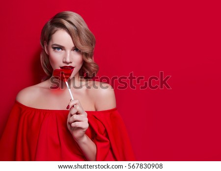 The girl in a red dress on a red background in the studio. Blonde girl holding a red heart-shaped lollipop. Valentine's Day. Advertising. Girl holding a lollipop in his mouth.