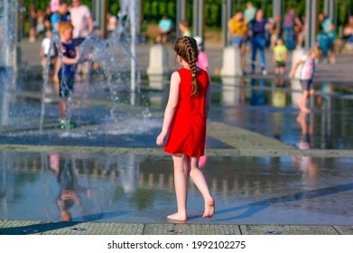 A girl in a red dress walks barefoot by the fountain on a hot sunny summer day in the city. Sultry heat on city streets, family vacation.
