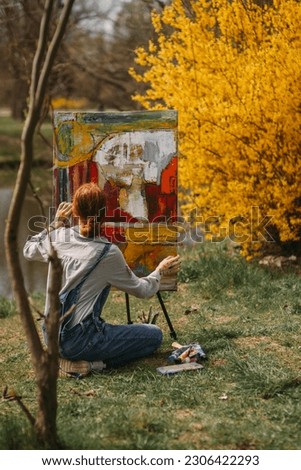 Girl with red hair and a bun painting a colorful picture on a big canvas while sitting on the gras near the lake
