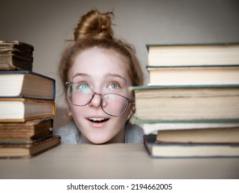 Girl With Red Hair In Glasses Peeps Between Two Stacks Of Books. Her Glasses Moved From Nose To Side.