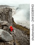 A girl in a red jacket and with a backpack sits on a rock and looks at the Dettifoss waterfall. Iceland