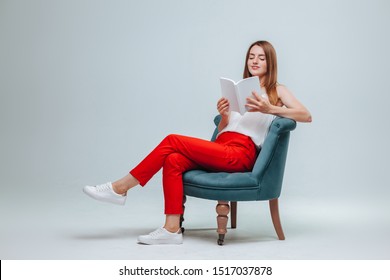 Girl In Red Pants Sitting In A Chair And Reading A Book With A White Cover