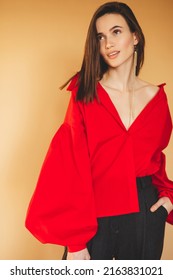 Girl in red shirt with puff sleeves, gold earrings and dark grey pants. Fashion studio portrait of young elegant woman wear minimalists outfit. Short hair brunette woman looks happy and laughing.