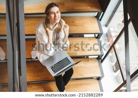 Girl rejoices in winning having raised her hands up sitting on stairs.Girl sitting in cafe, office or home make winner gesture.Woman blogger enjoys lot of views of her videos. View from above.