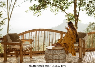 A girl is relaxing and drinking coffee on a balcony in a bamboo house overlooking the mountains. Coffee overlooking the mountains in Bali.