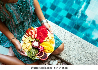 Girl Relaxing And Eating Fruit Plate By The Hotel Pool. Exotic Summer Diet. Photo Of Legs With Healthy Food By The Poolside. Tropical Beach Lifestyle.