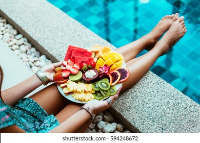 Girl Relaxing And Eating Fruit Plate By The Hotel Pool. Exotic Summer Diet. Photo Of Legs With Healthy Food By The Poolside. Tropical Beach Lifestyle.