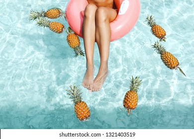 Girl Relaxing On Float Ring In Pool With Pineapples In Blue Clear Water, Photo Of Legs Closeup. Tropical Fruit Diet. Summer Holiday Idyllic Concept Top View.