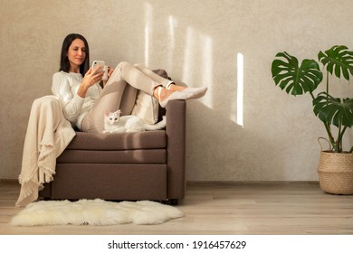 The girl is resting in a chair, drinking tea, reading a book, shopping on the phone. Shopping on the couch. The interior is minimalist. A white kitten is lying in a chair. - Powered by Shutterstock