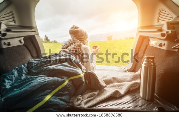 Girl resting in her car. Woman hiker, hiking\
backpacker traveler camper in sleeping bag, drinking hot tea and\
relaxing on top of mountain. Health care, authenticity, sense of\
balance and calmness.