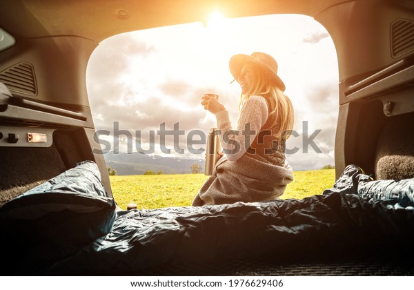 Girl resting in her car. Woman hiker, hiking\
backpacker traveler camper in sleeping bag, relaxing, drinking hot\
tea on top of mountain. Road trip. Health care, authenticity, sense\
of balance calmness.