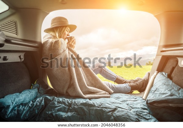 Girl resting in her car. Woman hiker, hiking\
backpacker traveler camper in sleeping bag, relaxing, drinking hot\
tea on top of mountain. Road trip. Health care, authenticity, sense\
of balance calmness.