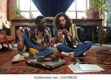 Girl With Retro Photocamera Showing It To Her Boyfriend While Relaxing On The Floor Of Living Room And Listening To Vynil Disks On Record Player