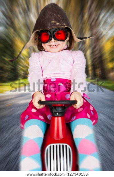 Girl in retro racing hat and goggles\
driving on toy car at speed with blurred\
background.