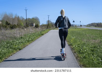 A girl rides an electric scooter along the road on a summer day, view from the back. - Shutterstock ID 2311787111