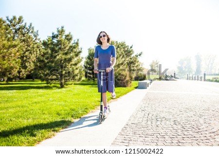 The girl rides a scooter in the park. Slim woman in dress have fun. Smiling brunette on a walk.