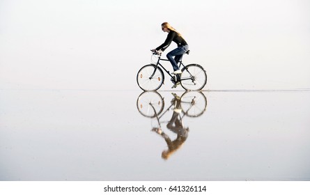 A Girl Riding A Bicycle By Water, That Creates A Bewitching Mirror Illusion.