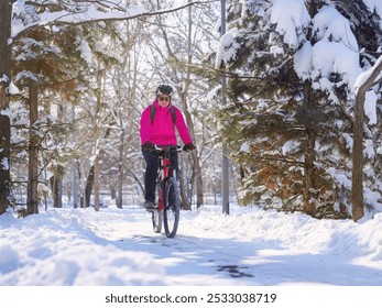 Girl riding a bike through a snowy park in winter on a sunny day. Winter cycling adventure. Girl in pink jacket and helmet on a bicycle - Powered by Shutterstock