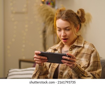 The girl in the room plays a video game on a portable game console. Interesting pastime, entertainment, mobile and video games, educational games. Close-up.