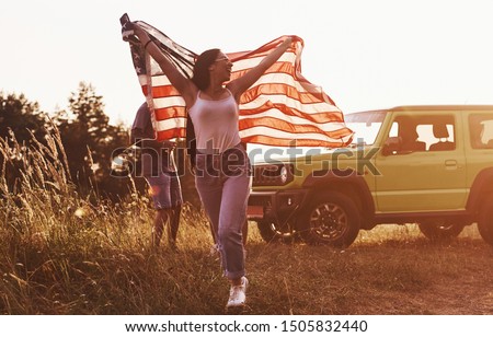 Girl runs forward. Friends have nice weekend outdoors near theirs green car with USA flag.
