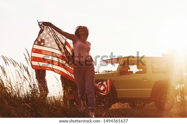 Girl runs forward. Friends have nice\
weekend outdoors near theirs green car with USA\
flag.