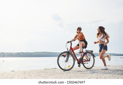 Girl Runs Near Bicycle. Two Female Friends On The Bike Have Fun At Beach Near The Lake.