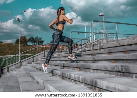 Girl runs in the summer in the city, on the morning run. Stair background, blue sky with clouds. Clothing leggings top. Free space for text. Phone earphones.