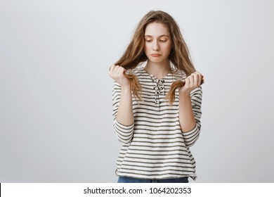 Girl Is Sad Of Her Bad Hair Strands. Portrait Of Gloomy Bothered Young Woman Holding Hair And Being Upset, Having Split Ends, Needing Help Of Hairdresser Or New Haircut, Standing Over Gray Wall