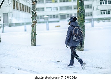 Girl Schoolgirl With A School Backpack Over Her Shoulders Runs To School. Child In The Background Of A School In Winter.