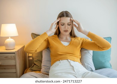 A girl is seated on a bed, holding her head with her hands. She appears deep in thought or experiencing distress. Her body language suggests emotional turmoil or physical discomfort. - Powered by Shutterstock