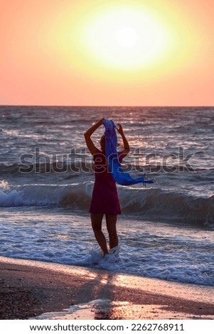 Girl with a shawl fluttering in the wind in her raised hand on the seashore at dawn, back view