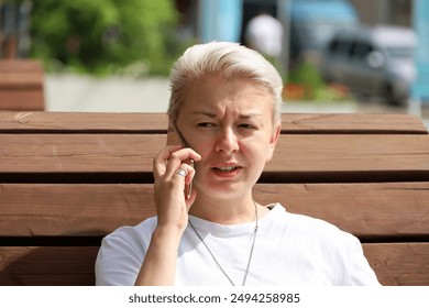 Girl with short blonde hair talking on mobile phone while sitting on street wooden bench. Tomboy lifestyle in summer city - Powered by Shutterstock