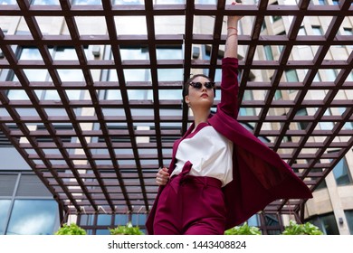 Girl With A Short Haircut, Glasses, Burgundy Jacket And Trousers Posing On The Street Near The Business Center. Women's Pantsuit Cherry Color.