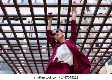 Girl With A Short Haircut, Glasses, Burgundy Jacket And Trousers Posing On The Street Near The Business Center. Women's Pantsuit Cherry Color.