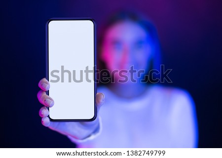 Girl showing blank smartphone screen over neon studio background, blue and pink lights