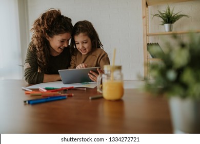 Girl Showing Her Mom New Games On Her Tablet. Sitting At Table, Taking A Break From Doing Homework.