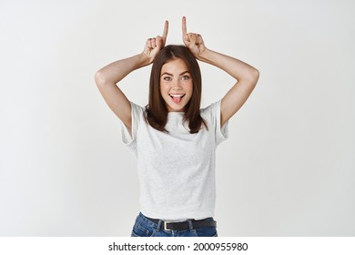 Girl Showing Inner Devil. Portrait Of Charming Cute And Confident European Female With Tattoo On Arm Holding Index Fingers Like Demon Horns On Head And Smiling With Intrigued Delighted Face