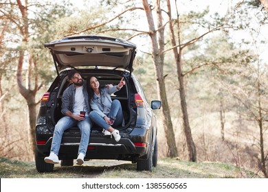 Girl shows next destination. Sitting on rear part of automobile. Enjoying the nature. Couple have arrived to the forest on their brand new black car.