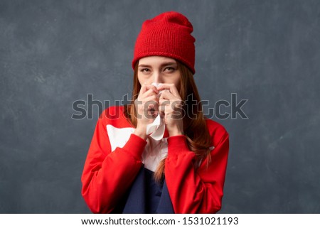 Girl sick, suffers runny nose, snot, feels unwell, sneezes into handkerchief, allergy. Woman with long redhead hair with bags under eyes in red hat, colored hoodie stands texture background in Studio