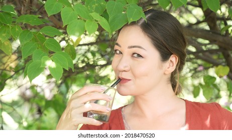 Girl Sipping Ice Coffee Through A Straw Of A Glass With Mean And Sneaky Eyes And Naughty Smile. Flirting Behaviour Of Asian Girl With White Skin. Idyllic Picnic Scene Portrait In A Beautiful Garden. 