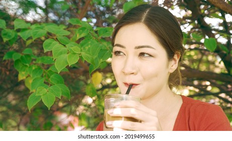 Girl Sipping Ice Coffee Through A Straw Of A Glass With Mean And Sneaky Eyes And Naughty Smile. Flirting Behaviour Of Asian Girl With White Skin. Idyllic Picnic Scene Portrait In A Beautiful Garden. 