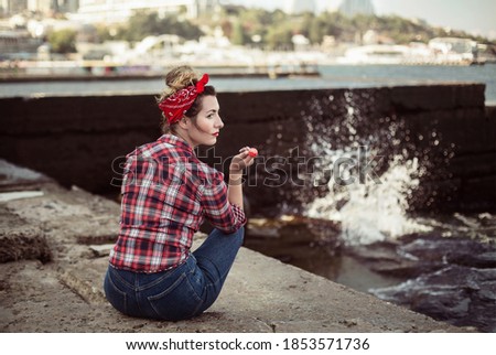 a girl sits by the sea in pin-up clothes and holds a candy on a stick in her hand