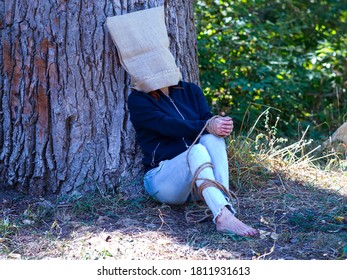 Ein Mädchen sitzt an einem Baum mit einer Tasche auf dem Kopf, ihre Hände sind mit einem Seil verbunden. Psychologie.