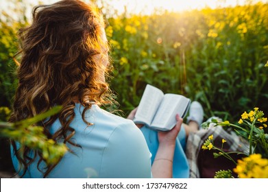 A Girl Sits In A Field On A Lawn Of Yellow Flowers, Reading A Book On A Summer Sunny Day. A Beautiful Girl In A Blue Dress Reads A Bible. Open Reading. Place For Text.